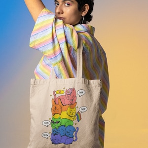 K&ouml;nnte beinhalten: Eine beige Tragetasche mit einer grafischen Illustration eines vertikalen Stapels aus f&uuml;nf bunten Katzen in Regenbogenfarben. Die Katzen sind rosa, orange, gelb, gr&uuml;n, blau und lila. Sprechblasen enthalten den Text They, Them, Their und Meow.