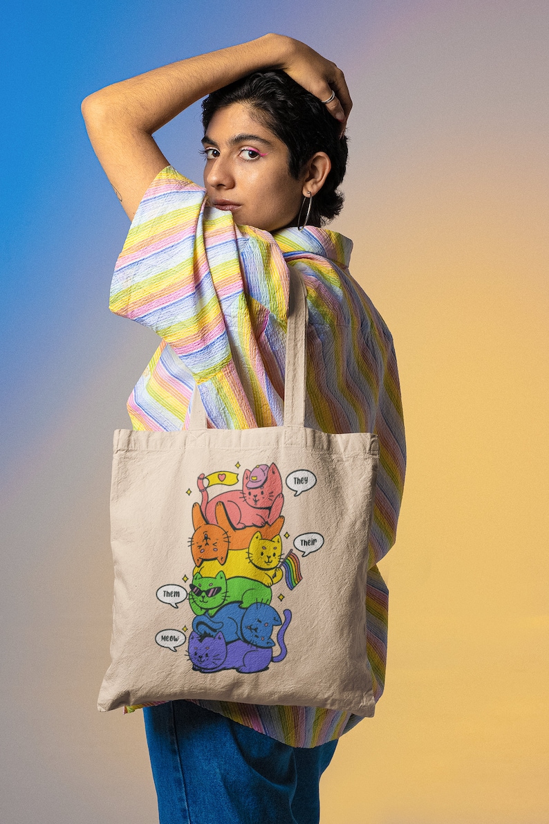 K&ouml;nnte beinhalten: Eine beige Tragetasche mit einer grafischen Illustration eines vertikalen Stapels aus f&uuml;nf bunten Katzen in Regenbogenfarben. Die Katzen sind rosa, orange, gelb, gr&uuml;n, blau und lila. Sprechblasen enthalten den Text They, Them, Their und Meow.