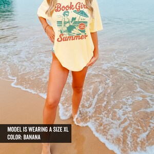 K&ouml;nnte beinhalten: Ein gelbes T-Shirt mit einer Retro-Grafik einer Frau, die am Strand ein Buch liest. Der Text auf dem Shirt lautet "Book Girl Summer" und "Guilty Pleasure Summer Reading".
