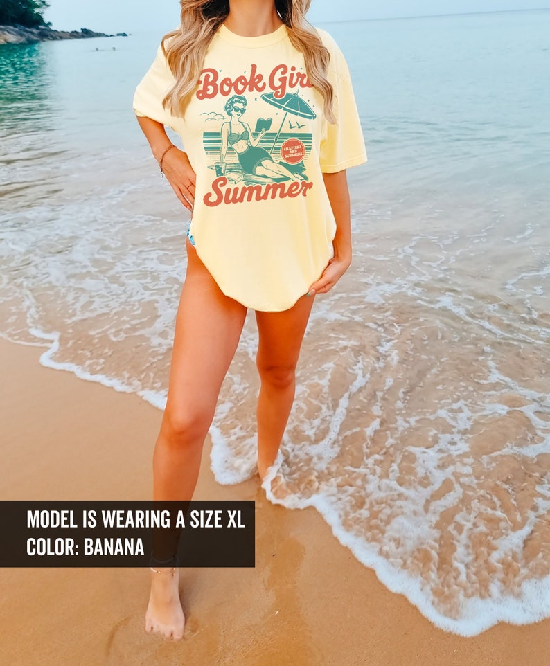 K&ouml;nnte beinhalten: Ein gelbes T-Shirt mit einer Retro-Grafik einer Frau, die am Strand ein Buch liest. Der Text auf dem Shirt lautet "Book Girl Summer" und "Guilty Pleasure Summer Reading".