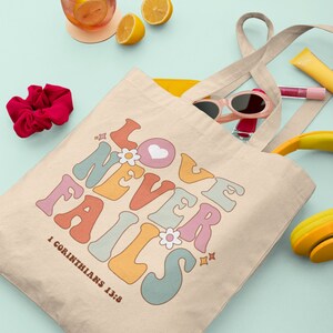 May include: A beige canvas tote bag with the text "LOVE NEVER FAILS" in colorful retro-style lettering. The text is surrounded by small flowers and stars. The bag has a yellow interior and two long beige straps. The text "1 Corinthians 13:8" is printed below the main text.