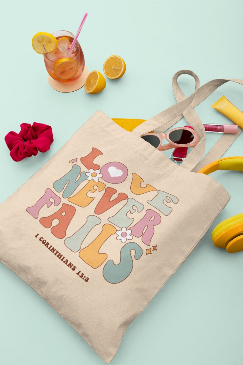 May include: A beige canvas tote bag with the text "LOVE NEVER FAILS" in colorful retro-style lettering. The text is surrounded by small flowers and stars. The bag has a yellow interior and two long beige straps. The text "1 Corinthians 13:8" is printed below the main text.