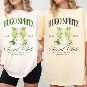 May include: Two white t-shirts with a graphic design of two glasses of Hugo Spritz, the text "Hugo Spritz", "Mar 2025", and "Social Club Emma's Bachelorette Washington, D.C.". The t-shirt on the left is ivory and the t-shirt on the right is banana yellow.