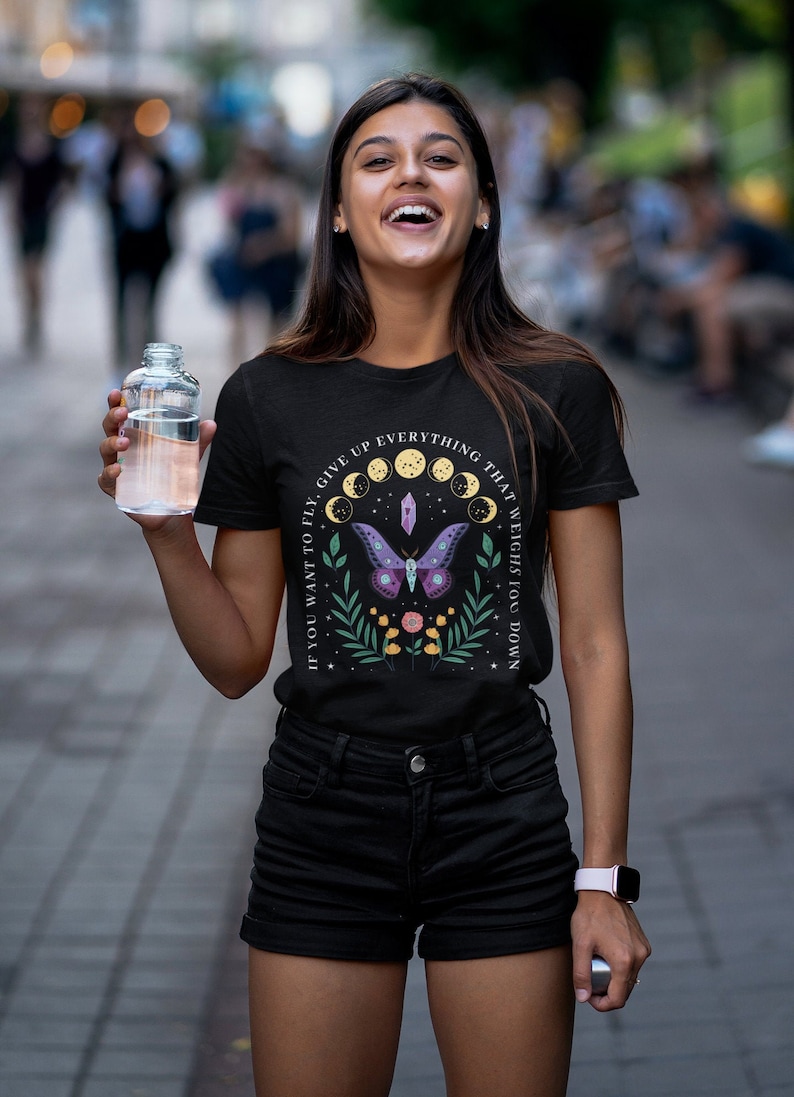 May include: Black t-shirt with a graphic of a purple butterfly, moon phases, and crystals. The text on the shirt reads "If you want to fly, give up everything that weighs you down."