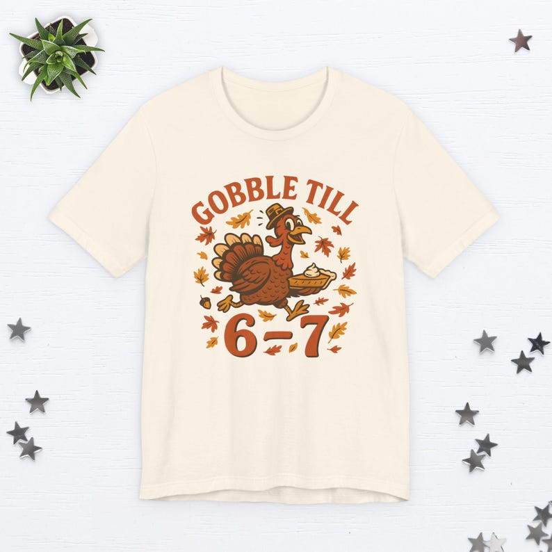 K&ouml;nnte beinhalten: Cremefarbenes T-Shirt mit einer Cartoon-Truthahn-Illustration, der einen Hut tr&auml;gt und einen Kuchen tr&auml;gt. Der Text "GOBBLE TILL" steht &uuml;ber dem Truthahn und "6-7" darunter. Herbstbl&auml;tter umgeben den Truthahn und den Text.