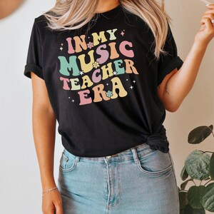 以下が含まれることがあります： レトロなフォントでカラフルなアクセントと花がついた「In My Music Teacher Era」という文字が書かれた黒いTシャツです。