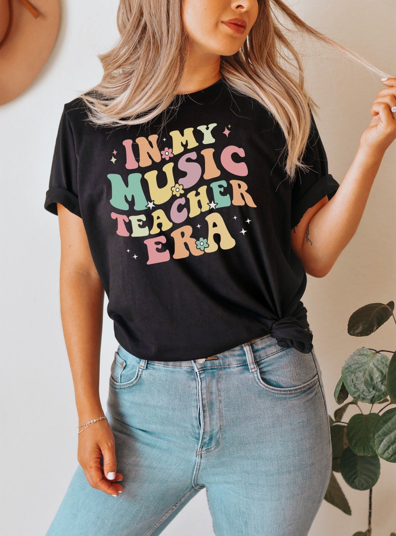 以下が含まれることがあります： レトロなフォントでカラフルなアクセントと花がついた「In My Music Teacher Era」という文字が書かれた黒いTシャツです。
