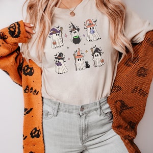 May include: A white t-shirt with a graphic of seven cute ghosts wearing witch hats and holding various Halloween items. The ghosts are surrounded by bats and stars.