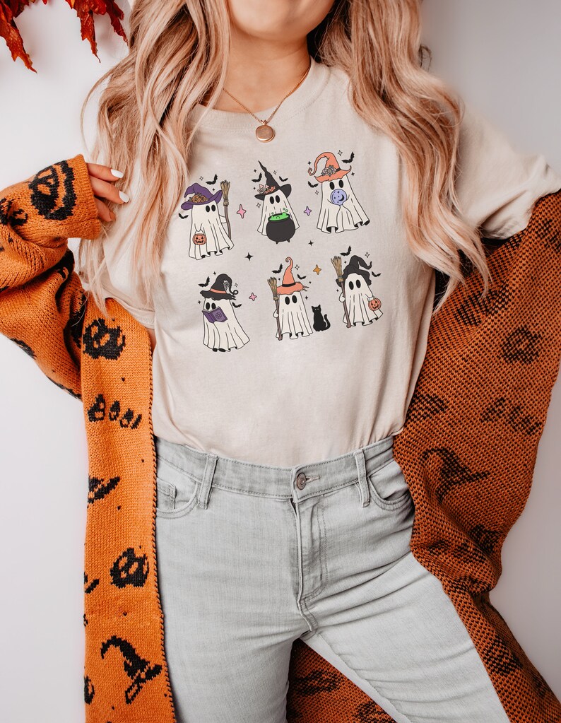 May include: A white t-shirt with a graphic of seven cute ghosts wearing witch hats and holding various Halloween items. The ghosts are surrounded by bats and stars.