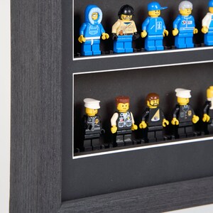 Puede incluir: Una caja de sombra negra con un paspart&uacute; blanco y dos filas de figuras Lego en miniatura. La fila superior presenta una figura con un abrigo de invierno azul, una figura con un casco marr&oacute;n y una chaqueta de color beige, una figura con una chaqueta azul y blanca, una figura con un uniforme azul y blanco, y una figura con un uniforme blanco. La fila inferior presenta una figura con un uniforme negro y un sombrero blanco, una figura con un chaleco negro y un sombrero marr&oacute;n, una figura con un uniforme negro y un sombrero negro, una figura con un uniforme negro y un sombrero amarillo, y una figura con un uniforme negro y un sombrero blanco.