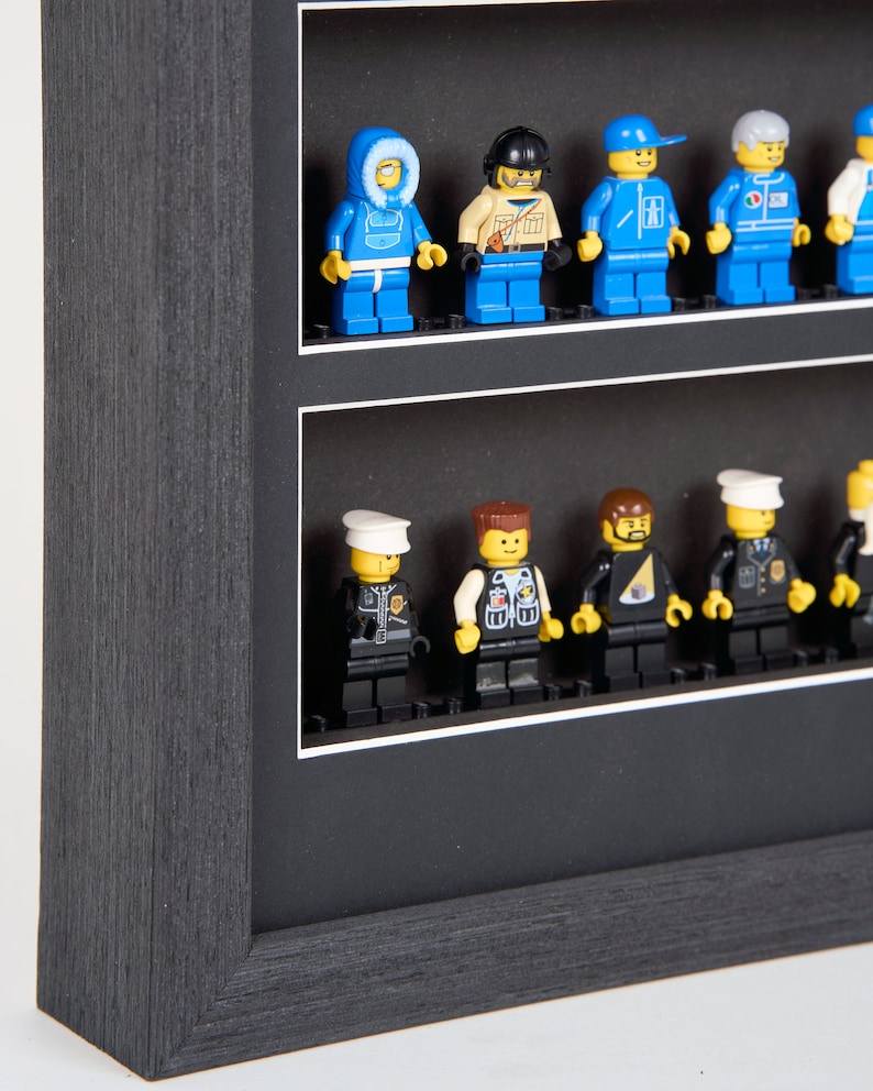Puede incluir: Una caja de sombra negra con un paspart&uacute; blanco y dos filas de figuras Lego en miniatura. La fila superior presenta una figura con un abrigo de invierno azul, una figura con un casco marr&oacute;n y una chaqueta de color beige, una figura con una chaqueta azul y blanca, una figura con un uniforme azul y blanco, y una figura con un uniforme blanco. La fila inferior presenta una figura con un uniforme negro y un sombrero blanco, una figura con un chaleco negro y un sombrero marr&oacute;n, una figura con un uniforme negro y un sombrero negro, una figura con un uniforme negro y un sombrero amarillo, y una figura con un uniforme negro y un sombrero blanco.