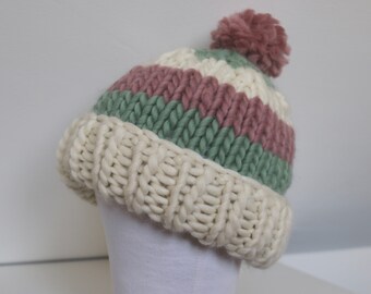 Puede incluir: Un gorro de punto blanco, rosa y verde a rayas con un pompón rosa esponjoso en la parte superior.
