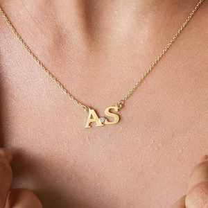 May include: A gold necklace featuring the initials "A" and "S" linked by a fine chain. A tiny, clear gemstone sits between the letters. The necklace is worn around the neck.