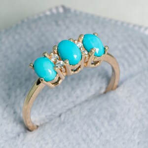 Puede incluir: Un anillo de oro con tres piedras turquesas ovaladas. Pequeñas piedras claras están engastadas entre las piedras turquesas. El anillo está sobre una superficie de terciopelo gris.