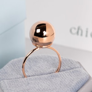 May include: A rose gold ring with a large, faceted sphere as its centerpiece. The ring is displayed on a gray velvet surface, with the brand name "chicvida" visible in the background. The ring's design is minimalist and modern.
