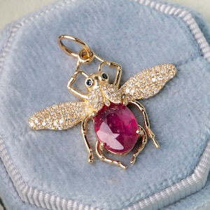 May include: A gold-coloured bee pendant featuring a large oval ruby gemstone. The wings are adorned with small, clear gemstones. Two small blue gemstones are set above the ruby. The pendant is presented in a light blue velvet jewellery box.