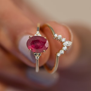 Può includere: Un anello d'oro con una grande pietra di rubino ovale circondata da diamanti più piccoli. Un secondo anello d'oro con una fila di diamanti a forma di V si trova sotto il primo anello.