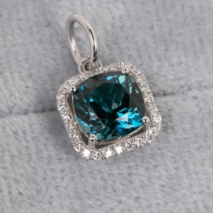 May include: A square-shaped pendant with a large, dark blue gemstone surrounded by a halo of smaller, white gemstones. The pendant is made of silver and has a loop at the top for attaching to a chain.