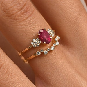 Könnte beinhalten: Ein goldener Ringsatz mit einem großen ovalen Rubin-Edelstein und kleineren Akzentdiamanten. Der Ring ist mit einem gebogenen Band aus kleinen Diamanten kombiniert. Der Schmuck wird am Finger einer Person präsentiert.