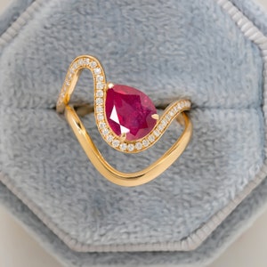 May include: A gold ring with a pear-shaped ruby gemstone surrounded by a halo of smaller diamonds. The ring has a unique, wave-like design.