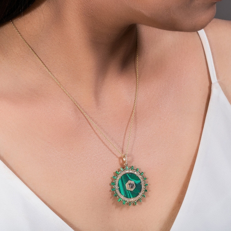 May include: A gold necklace with a large pendant featuring a circular malachite stone center. The pendant is surrounded by a halo of green gemstones and small diamonds. The necklace is worn against a white top.