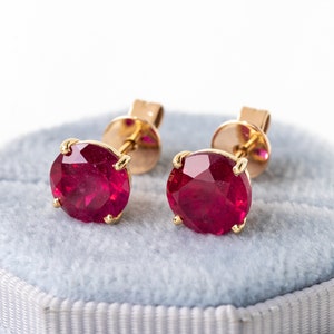 May include: A pair of ruby stud earrings. Each earring has a round, faceted, deep red gemstone set in gold-coloured metal. The earrings are displayed on a light blue velvet surface.