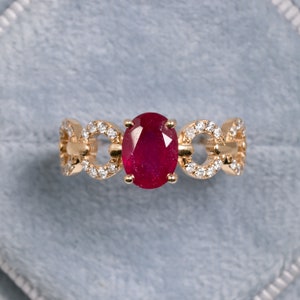 以下が含まれることがあります： 大きな楕円形のルビーの宝石を、小さなダイヤモンドで囲んだ金の指輪。繊細な連結デザインでセットされています。