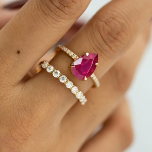 May include: Three gold rings are shown. One ring has a large, pear-shaped, ruby-red gemstone. Another ring features a row of small, round, clear gemstones. The third ring is a thin band with small, clear gemstones.