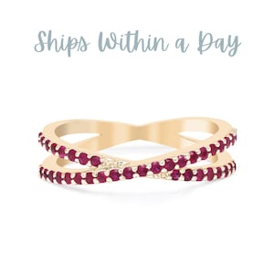 May include: A gold-colored ring with a crisscross design, adorned with small, vibrant red gemstones. The ring has a smooth, polished finish and is set against a white background. The text "Ships Within a Day" is above the ring.