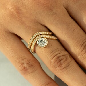 May include: A gold ring with a unique design featuring a large, round, clear gemstone in the center. The ring has two curved bands, each lined with small, sparkling gemstones. The ring is on a person's finger.