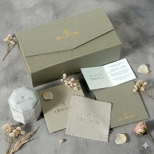 May include: An assortment of jewelry packaging and accessories in a soft, muted color palette. Includes a sage green box, a light blue octagonal ring box, cards, and a cleaning cloth, all branded with "chicvida" in gold.