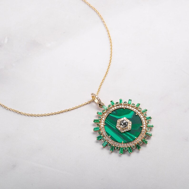 May include: Gold-toned necklace with a large, round pendant. The pendant features a vibrant green malachite center, surrounded by emerald-cut green stones and small clear stones. A small, clear stone is set in the center of the malachite.