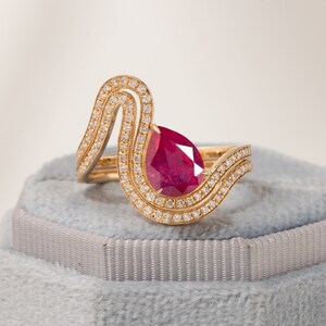 Könnte beinhalten: Ein goldener Ring mit einem birnenförmigen Rubin, der von einem Heiligenschein aus kleineren Diamanten umgeben ist. Der Ringarm ist ein Doppelarm mit einem wellenförmigen Design.