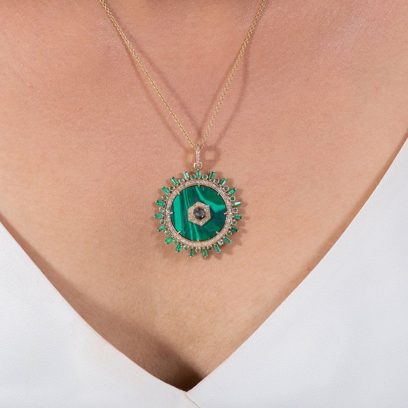 May include: A gold necklace featuring a large, circular pendant. The pendant has a vibrant green malachite center with a dark gray hexagonal stone. The pendant is surrounded by emerald-cut green stones and small diamonds, with a delicate gold chain.