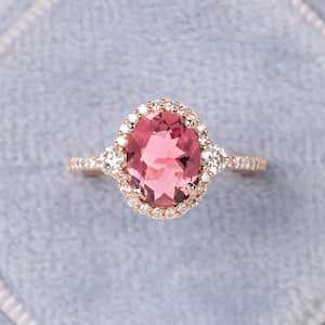 May include: Rose gold ring featuring a large oval pink gemstone surrounded by a halo of smaller white gemstones. Additional white gemstones flank the central stone on either side. The ring band is also adorned with small white gemstones.