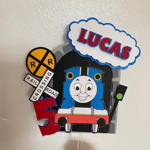 Op de afbeelding: Een blauwe en rode Thomas de Tank Engine taarttopper met een witte wolk waarop "LUCAS" staat. De locomotief staat voor een grijze tunnel met een geel en zwart spoorwegovergangsbord en een rood nummer 5.