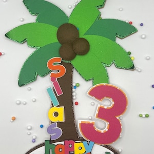 May include: A green palm tree cake topper with brown coconuts, a colorful "Happy Birthday" banner, and a pink number "3" with white polka dots.