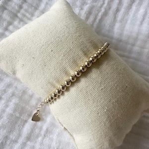Peut inclure: Un bracelet en perles dorées avec un petit charme en forme de cœur. Le bracelet est sur un fond en tissu blanc.