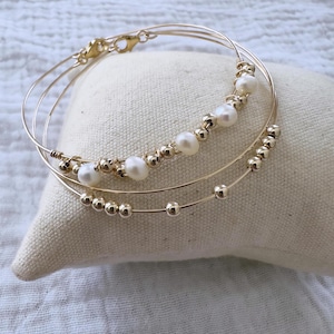 Peut inclure: Trois bracelets en fil d'or. L'un est orné de perles blanches, un autre de perles dorées, et le troisième est uni. Les bracelets reposent sur un coussin beige.