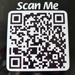 May include: A white square with rounded corners featuring a black and white QR code. The text "Scan Me" is printed in white at the top of the square. The QR code is a complex pattern of black squares and rectangles.