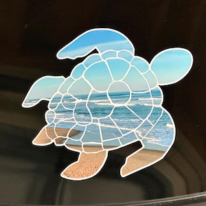 May include: A turtle-shaped sticker with a mosaic design. The mosaic depicts a beach scene with blue skies, ocean waves, and sandy shores. The sticker has a white outline and is set against a black background.