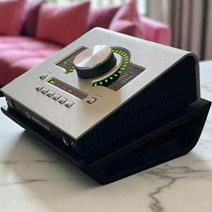 May include: Silver and black audio interface device with a large control knob and multiple buttons. The device is on a black stand, and the background includes a pink sofa and a marble table.