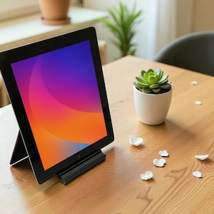 以下が含まれることがあります： 黒いタブレット。オレンジ、紫、青の鮮やかな画面表示で、黒いスタンドに立てかけられています。白い鉢に入った小さな多肉植物と白い花びらが木製のテーブルの上にあります。