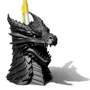 Peut inclure: Un porte-crayon en forme de tête de dragon noir mat avec des écailles texturées et des dents pointues. La tête de dragon présente une bouche ouverte et une section supérieure creuse contenant actuellement un stylo jaune et blanc unique.