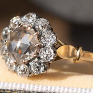 Może przedstawiać: Złoty pierścionek z dużym, fasetowanym diamentem otoczonym mniejszymi diamentami. Oprawa pierścionka jest złota, z otoczką mniejszych diamentów otaczających centralny kamień. Pierścionek jest umieszczony na miękkim, neutralnym tle.