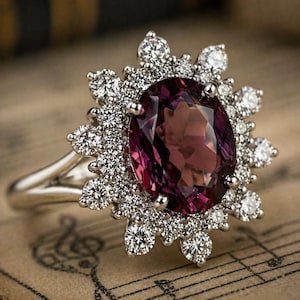 Puede incluir: Un anillo de plata con una gran gema ovalada de color rojo púrpura, rodeada de gemas redondas y transparentes más pequeñas. El anillo tiene una banda fina y pulida y un engaste intrincado. Se muestra sobre papel envejecido con notación musical.