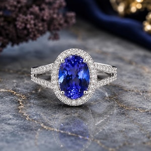 May include: A silver-toned ring featuring a large, oval-cut blue gemstone at the center, surrounded by a halo of small, clear, round-cut stones. The band is split into two rows on each side, both encrusted with small, clear, round-cut stones.