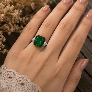 May include: A ring featuring a large, rectangular emerald-cut green gemstone set in yellow gold prongs, flanked by two triangular-cut clear white diamonds on each side, mounted on a thin, polished metal band.