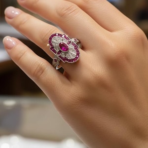 May include: A silver ring featuring a central oval ruby surrounded by smaller rubies and clear gemstones. The ring has an Art Deco design with a floral-like shape, and is worn on a finger.