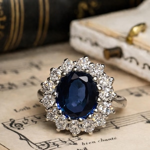 May include: A silver ring featuring a large, oval, deep blue gemstone surrounded by a halo of smaller, clear gemstones. The ring rests on a sheet of vintage sheet music with musical notes and the words "bien chanté".